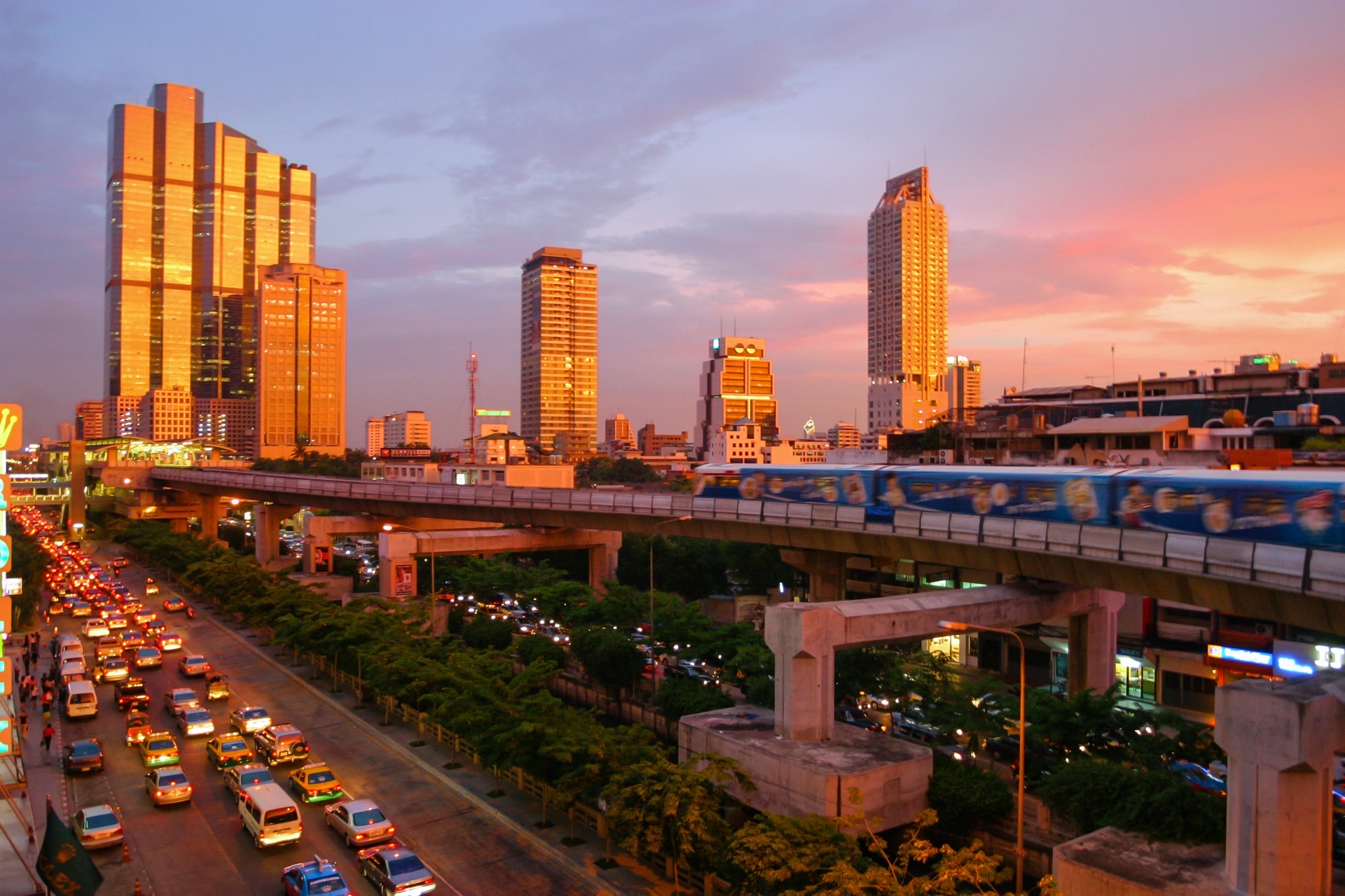 Bangkok BTS Skytrain navigating elevated rails over busy urban avenue during sunset surrounded by modern skyscrapers and digital nomad hubs
